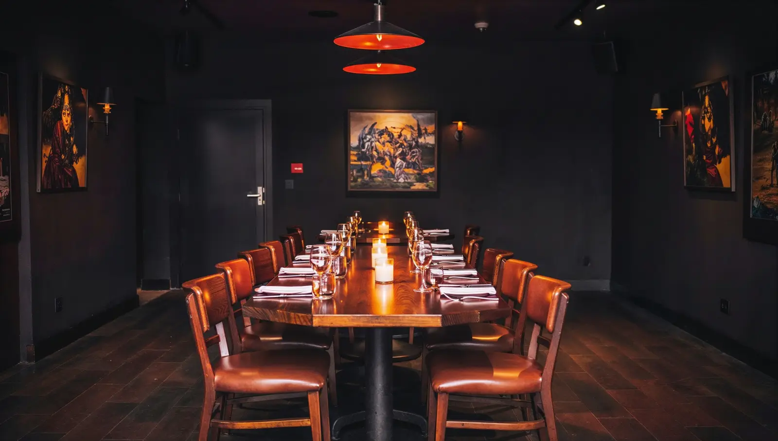 Private dining room with a long candlelit table set for a party, warm pendant light, framed art on dark walls.