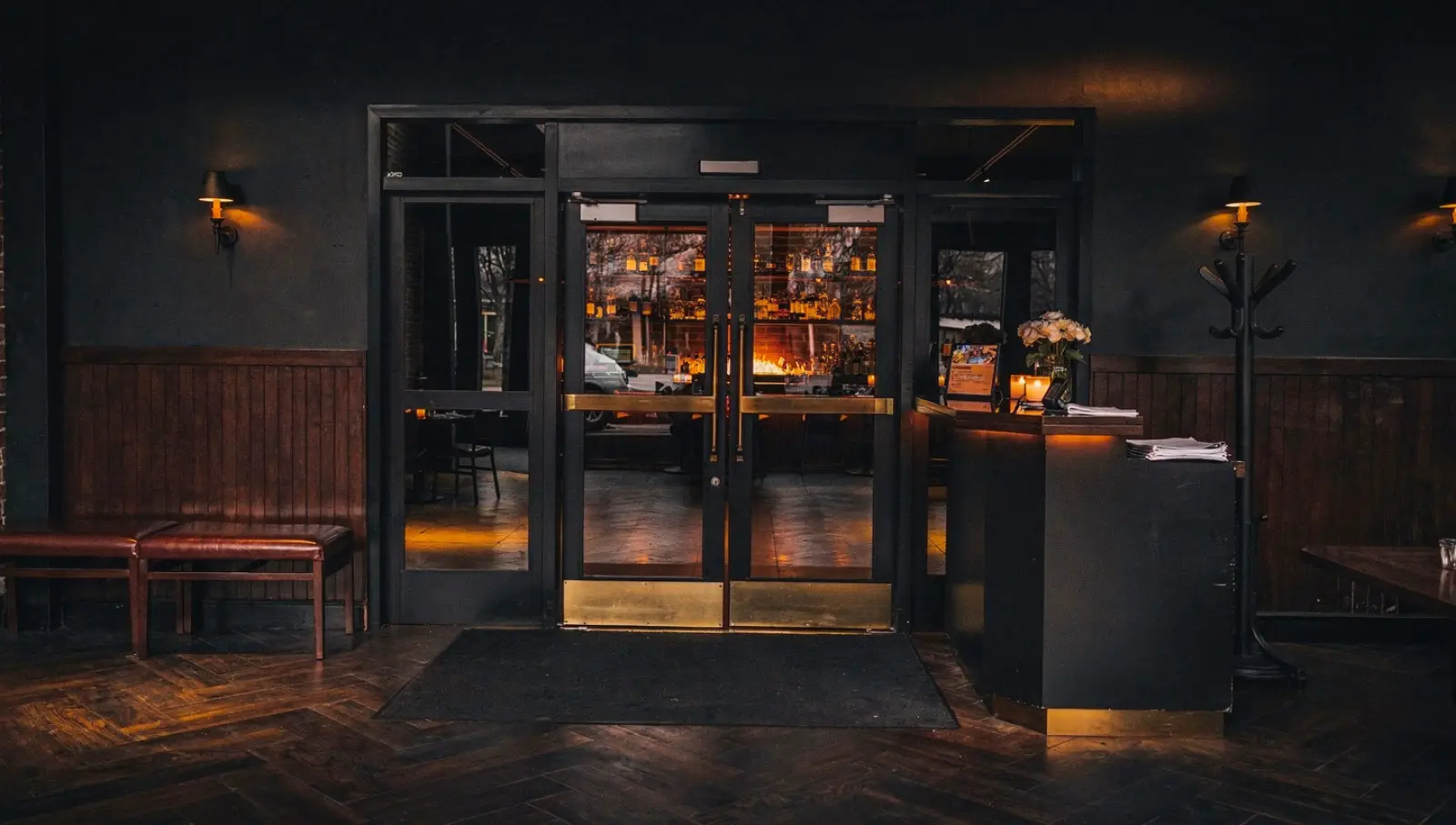 Dark timber-panelled entrance with brass-trimmed glass doors, a host stand with white roses, the bar glowing beyond.