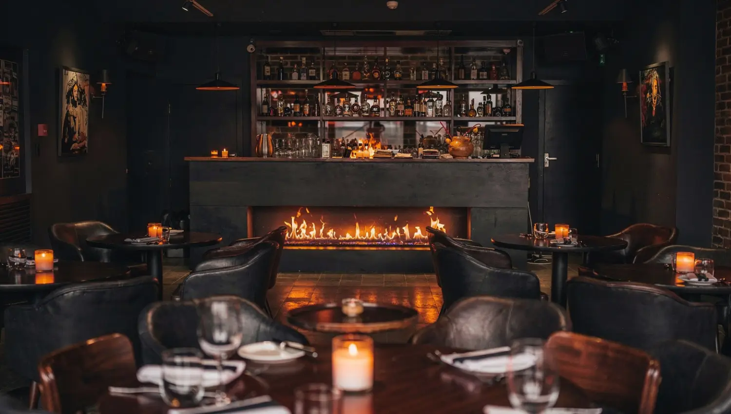 Low-lit lounge with leather tub chairs drawn around a long open fire, a spirits bar glowing behind.