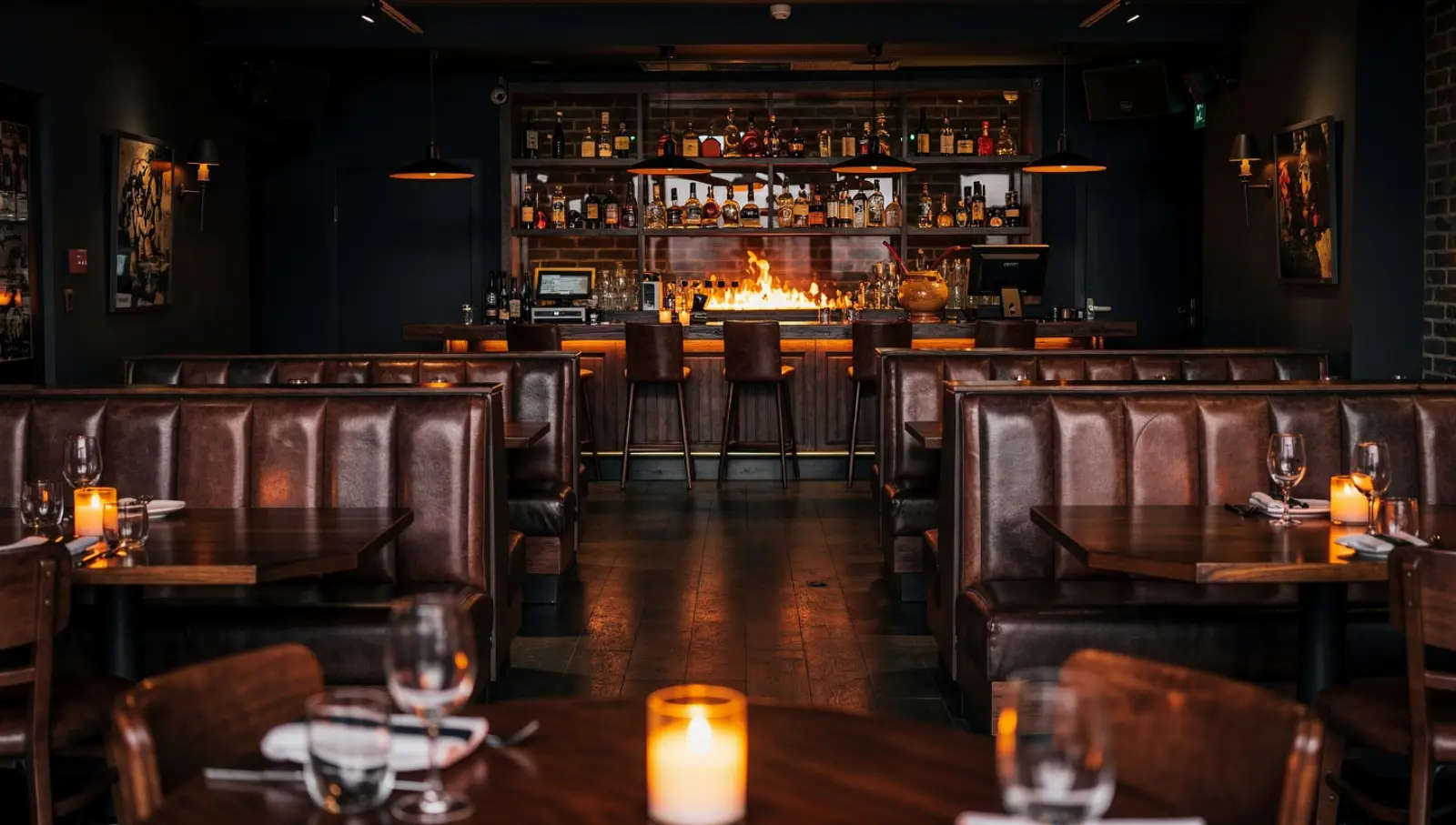 Public dining room with high-backed leather booths either side of a central aisle, candlelit tables, and a bar with an open flame and bottles behind.